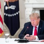 Donald Trump looks at a smartphone while sitting at a desk.