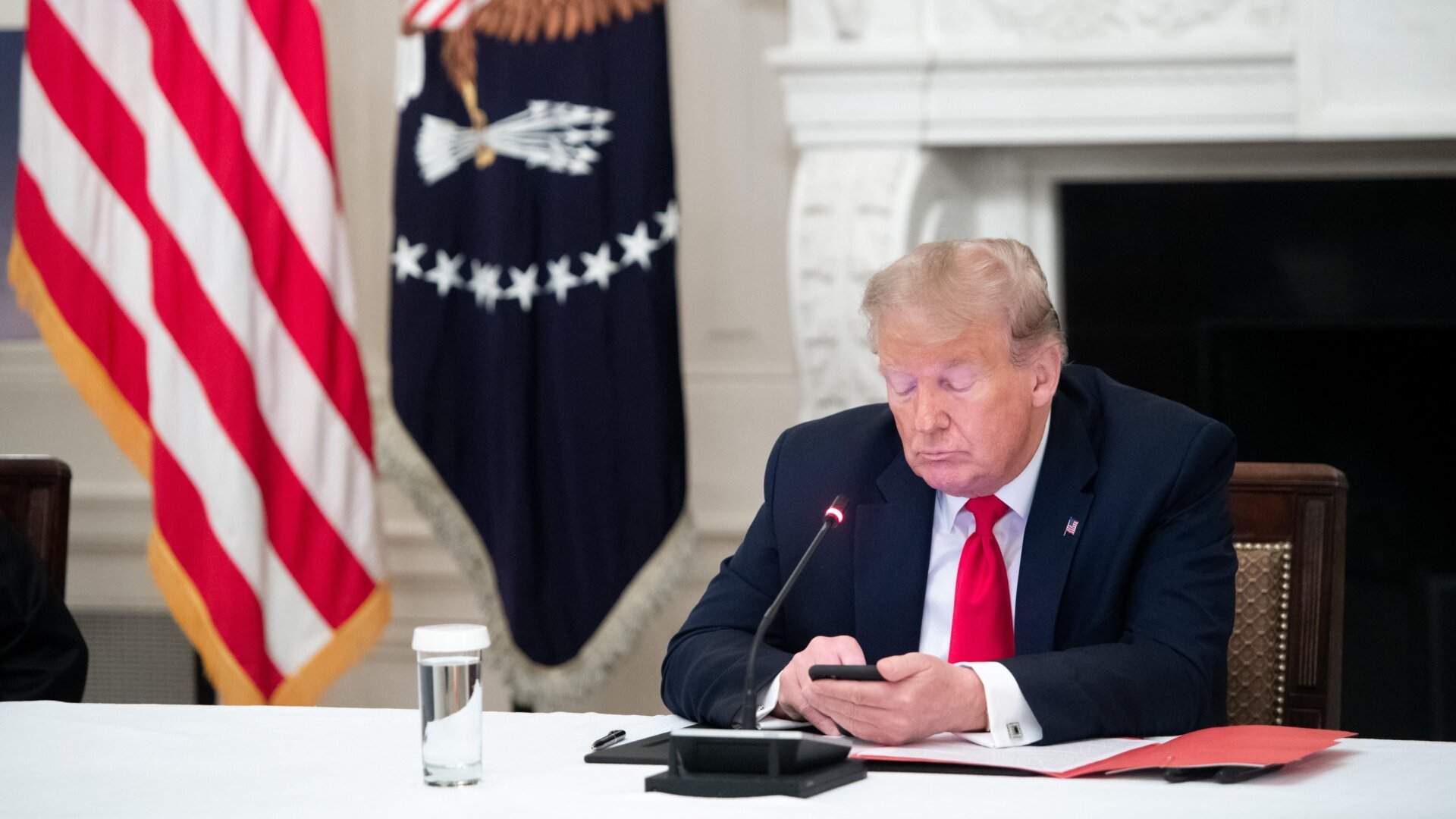Donald Trump looks at a smartphone while sitting at a desk.
